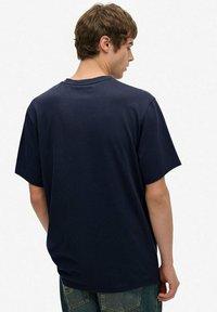 Jonge man met kort bruin haar, gekleed in een eenvoudige marineblauwe T-shirt en vervaagde blauwe spijkerbroek, staat zijwaarts met zijn hoofd iets naar achteren gedraaid.