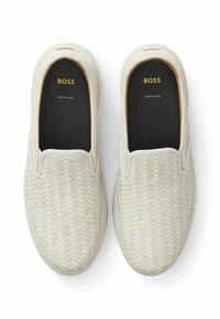 Slip-on-skor i vävt, ljuskrämigt material med en texturerad yta. Svart inredning med guldig märkning: "BOSS" och "CRAFTED IN ITALY."