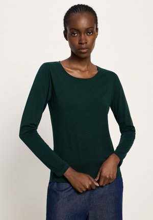 Woman with braided hair wearing a dark green long-sleeve shirt and blue jeans, standing with hands clasped at waist against plain background.