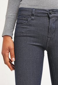 Jeans skinny de denim oscuro con diseño de cinco bolsillos, cierre frontal visible con botón y textura sutil, que complementan una camiseta de manga larga gris.
