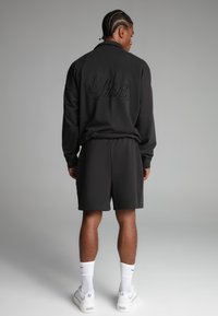 Muž s opletenými vlasmi stojí otočený chrbtom, oblečený v čiernom tričku s dlhým rukávom s výšivkou "Siksilk", zodpovedajúcich šortkách, bielych ponožkách a teniskách.