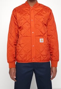 Quiltad orange jacka med rund krage, tryckknappar och sidofickor. Har en liten logoval med en gul grafisk detalj på den nedre framsidan.