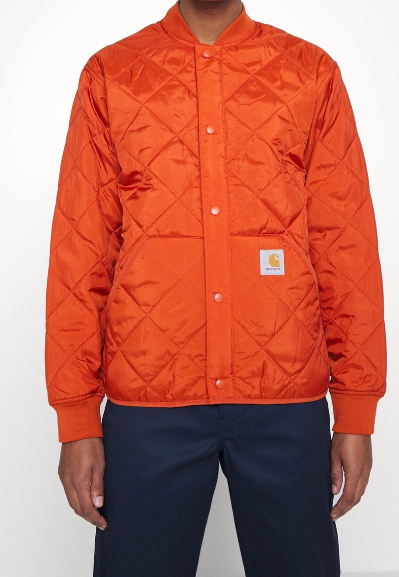 Quiltad orange jacka med rund krage, tryckknappar och sidofickor. Har en liten logoval med en gul grafisk detalj på den nedre framsidan.