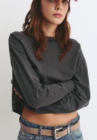 Maglietta a maniche lunghe in cotone grigio carbone con vestibilità rilassata, abbinata a jeans di denim blu a vita alta e una cintura beige con fibbia argentata.