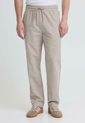 Man wearing beige drawstring pants and beige sneakers with white stripes, standing against a plain light background.