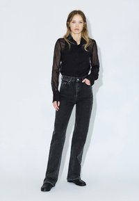 Blusa nera trasparente con chiusura a bottoni e maniche lunghe, abbinata a jeans neri a vita alta. Il modello si trova su uno sfondo chiaro.