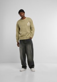 Strickpullover in hellem Olivgrün mit strukturiertem Gewebe und einem weißen Emblem auf der Brust, kombiniert mit übergroßen grauen Jeans und weißen Sneakers.