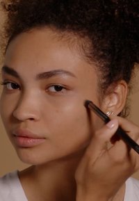 Makeup brush applying highlighter on a smooth, light brown skin tone. Subject has curly hair and is wearing a simple white top.
