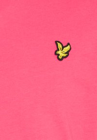 T-shirt en coton rose avec un petit logo d'aigle noir et or brodé sur la poitrine, fabriqué avec un tissu lisse et texturé.