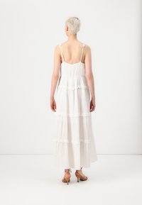 Hvit maxikjole med lagdelt stoff og blonderkant, justerbare spaghetti stropper og løs passform. Brukt med beige hæl sandaler.