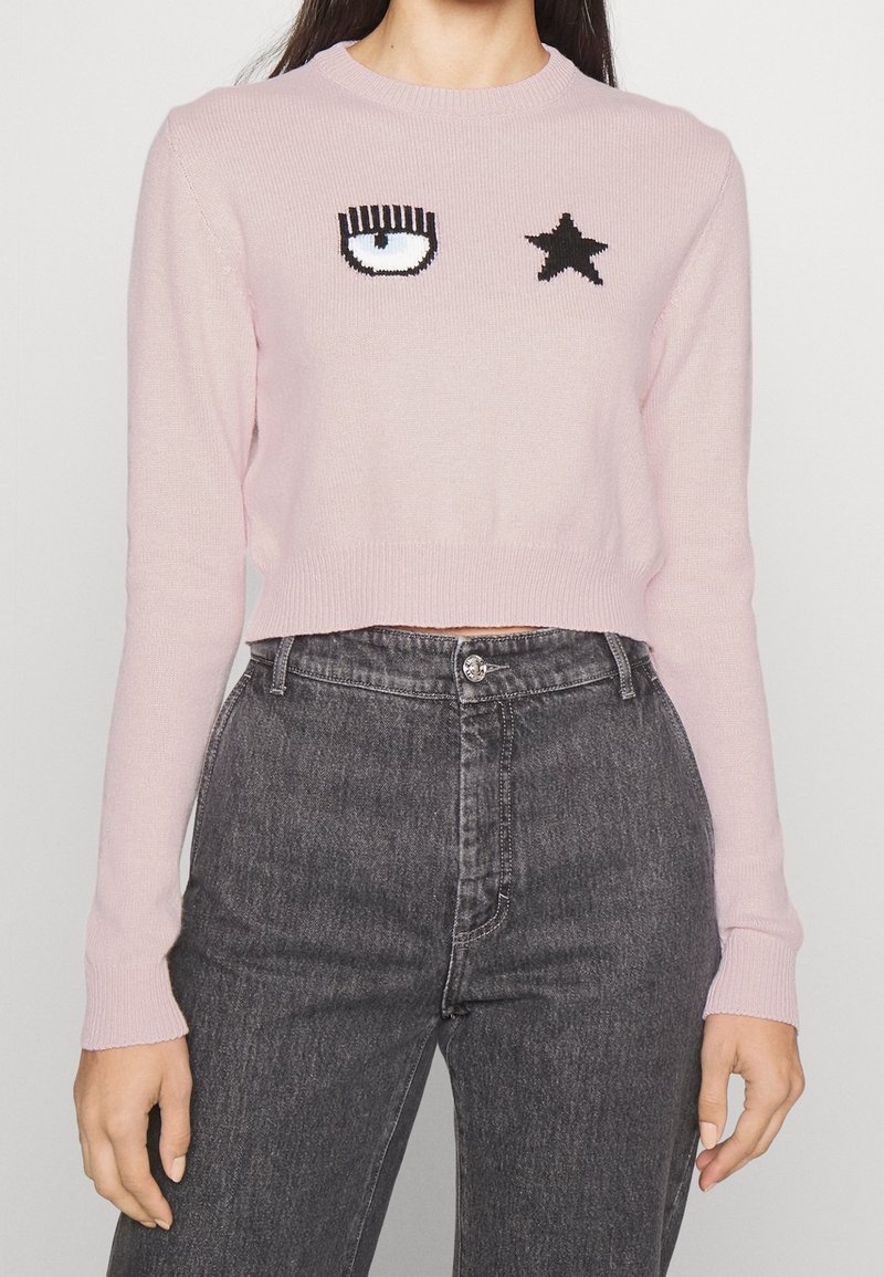 Femme portant un pull court rose clair avec un motif œil unique et étoile, associé à un jean gris foncé taille haute.