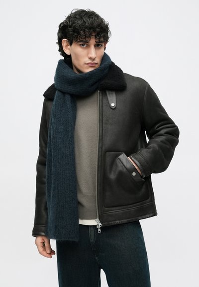 Juoda odinė striukė su kailio apykakle, po ja pilkas megztinis, tamsiai mėlynas šalikas. Matoma šoninė kišenė ir metalinės detalės.