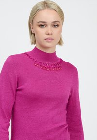 Giovane donna con capelli corti biondi che indossa un maglione rosa brillante decorato con perline rosa lungo scollatura.