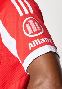 Rød atletisk drakt med hvite detaljer og tre vertikale striper. Har en sirkulær logo med en QR-kode og "Allianz" i fet skrift.