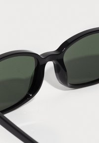 Gafas de sol negras con montura redondeada y lentes verde oscuro. La montura tiene un acabado brillante, presenta una textura suave y no tiene marcas visibles.