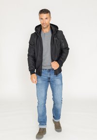 Czarna bomberka z kapturem i zapinanymi kieszeniami, noszona na szarym t-shircie, w połączeniu z niebieskimi dżinsami i oliwkowymi butami.