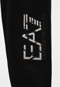 Čierne látkové nohavice s textúrovaným strieborným logom a geometrickým vzorom na ľavej nohe. Zobrazený je tiež názov značky "Emporio Armani".