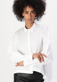 Camicia bianca a maniche lunghe con colletto a punta, chiusura con bottoni e vestibilità rilassata. Realizzata in tessuto liscio; presenta polsini larghi e orlo curvo.