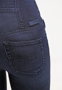 Gros plan sur la poche arrière d'un jean en denim bleu foncé avec un détail de couture et une petite étiquette rectangulaire de la marque Diesel au-dessus de la poche.