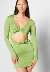 Vestido mini verde de mangas longas com um decote em V profundo, recorte em forma de olho-chave no peito e textura lisa e brilhante. A silhueta justa acentua as curvas.