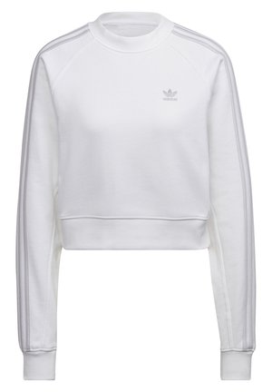Vrouw draagt een wit crop-top sweatshirt met lange mouwen en drie strepen op de mouwen, gecombineerd met roze trainingsbroek met witte zijstrepen.