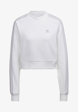 Vrouw draagt een wit crop-top sweatshirt met lange mouwen en drie strepen op de mouwen, gecombineerd met roze trainingsbroek met witte zijstrepen.
