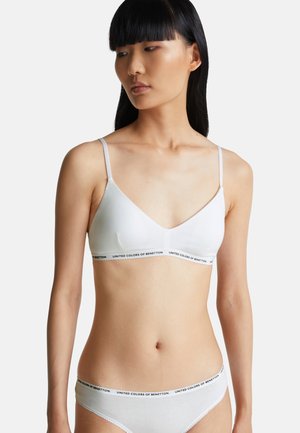 Vrouw met lang zwart haar die een witte beha en bijpassend ondergoed draagt met de tekstband "United Colors of Benetton" op een neutrale achtergrond.