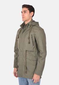 Parka giacca verde oliva con cappuccio, dettagli con cordoncino, tasche laterali e tasca sul petto con zip. Realizzata in un tessuto durevole e testurizzato.