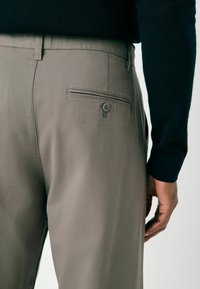 Gray trousers made from a smooth fabric, featuring a decorative back pocket with a button and a fitted cut, paired with a dark sweater.