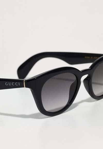 Ochelari de soare Gucci negri cu lentile rotunde, cu un gradient, și rame lucioase, pe un fundal alb.