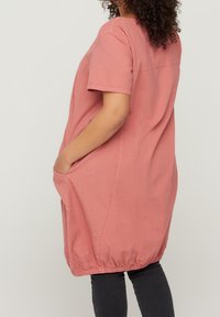 En kvinna står på sidan, iförd en kortärmad rosa klänning med sidofickor och en volang nederkant, tillsammans med mörka jeans.