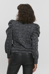 Sweater med puffærmer i mørkegrå med et struktureret zigzag-mønster, der har en tætsiddende kant og et blødt, strækbart stof. Sammen med sorte bukser.