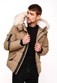 Beige puffjacka med avtagbar huva i päls, fickor med dragkedja och en snug passform. Funktioner svarta accenter och en slät yta.