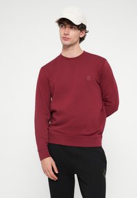 Bordó pulóver kerek nyakkal, rugalmas passzékkal és márkajelzéssel ellátott folttal. Fekete nadrággal párosítva, a modell egy "BOSS" logós világos sapkát visel.