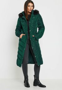 Cappotto puffer verde lungo con cappuccio bordato in pelliccia, texture trapuntata, chiusura a bottoni e zip, e due tasche laterali. Indossato sopra jeans neri e stivali.