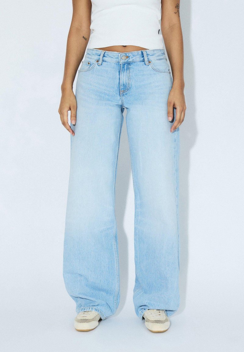 Light blue wide-leg jeans made from denim, featuring a mid-rise waist, five pockets, and subtle fading. Paired with light-colored sneakers.