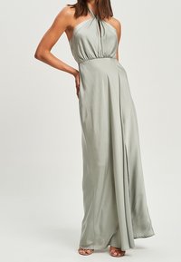 Frau in einem bodenlangen, halterlosen, hellgrünen Satin-Kleid mit einem Schlitz vorne, steht mit einer Hand auf der Hüfte.