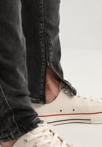 Pantalones de mezclilla negra con cremalleras laterales, que exponen la piel y zapatillas de lona de color claro con acentos rojos y azules.