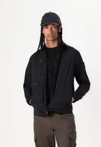 Black lightweight jacket with a zip front, one chest pocket, and a relaxed fit, paired with a black cap and dark cargo pants.