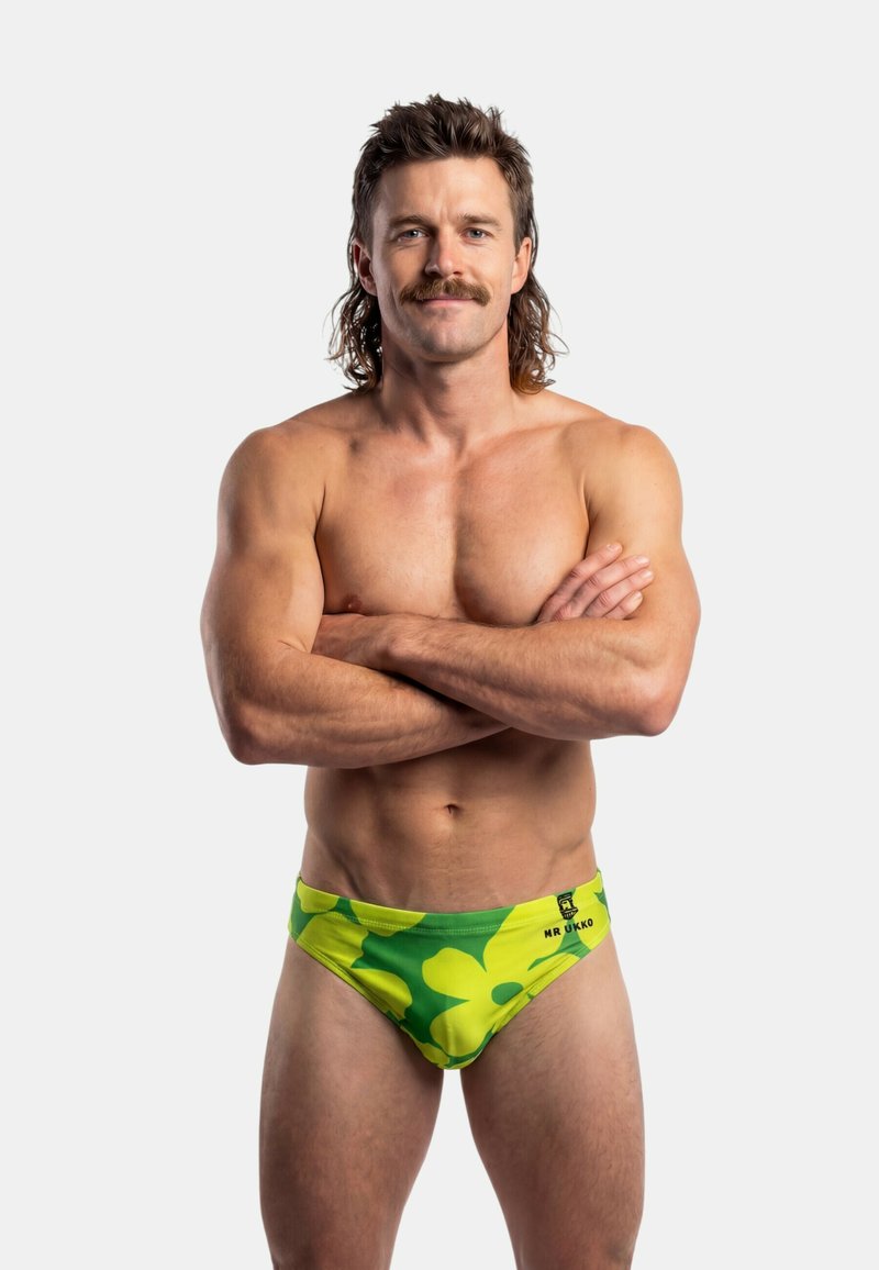 Hombre musculoso con mullet y bigote, de pie con los brazos cruzados, vistiendo un bañador floral verde y amarillo contra un fondo claro y liso.