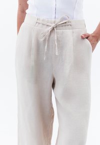 Pantalons en lin beige avec une ceinture à cordon, dotés de poches latérales et d'une coupe décontractée et ample, mettant en valeur un tissu subtilement texturé.