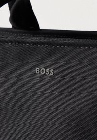 Μαύρη υφασμάτινη τσάντα με υφή στην επιφάνεια, εμφανείς ραφές και εμφανές γκρι λογότυπο «BOSS». Απλός σχεδιασμός χωρίς επιπλέον διακοσμητικά στοιχεία.