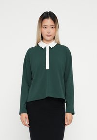 Mörkgrön långärmad polo topp med vit krage och knapplist, tillverkad av mjukt tyg med en avslappnad passform.