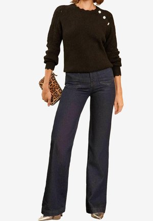 Kobieta ubrana w czarny dzianinowy sweter z trzema guzikami na ramieniu, ciemne, wysokowcześnie szerokie spodnie typu jeans, metaliczne buty w szpic, trzymająca kopertówkę w panterkę.