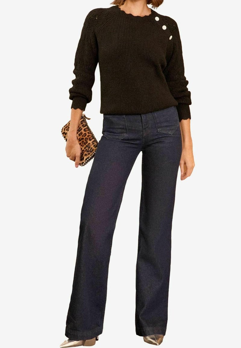 Femme portant un pull en maille noir avec trois boutons sur l'épaule, un jean large taille haute foncé, des chaussures pointues métalliques, tenant une pochette à imprimé léopard.