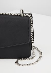 Borsa a tracolla nera con superficie testurizzata, tracolla in catena argentata e chiusura a patta. Un dettaglio in metallo argentato di rilievo arricchisce il design.