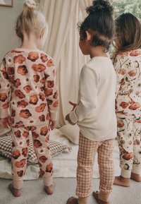 Tre børn i pyjamas: bluse og bukser med blomsterprint i ferskentoner, ensfarvet cremefarvet bluse og ternede tanbukser med manchetter.