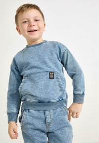 Niebieski dresowy sweter w stylu dżinsowym z kieszenią z przodu i logo, w połączeniu z pasującymi dżinsami. Luźny krój z ściągaczami na rękawach i dole.