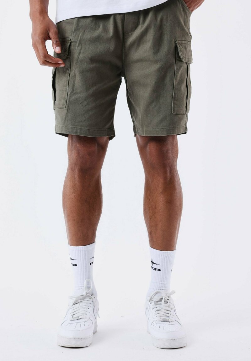 Pantalones cortos de cargo verde oliva con múltiples bolsillos, hechos de tela resistente. Se llevan con calcetas blancas y zapatillas deportivas para un look casual.