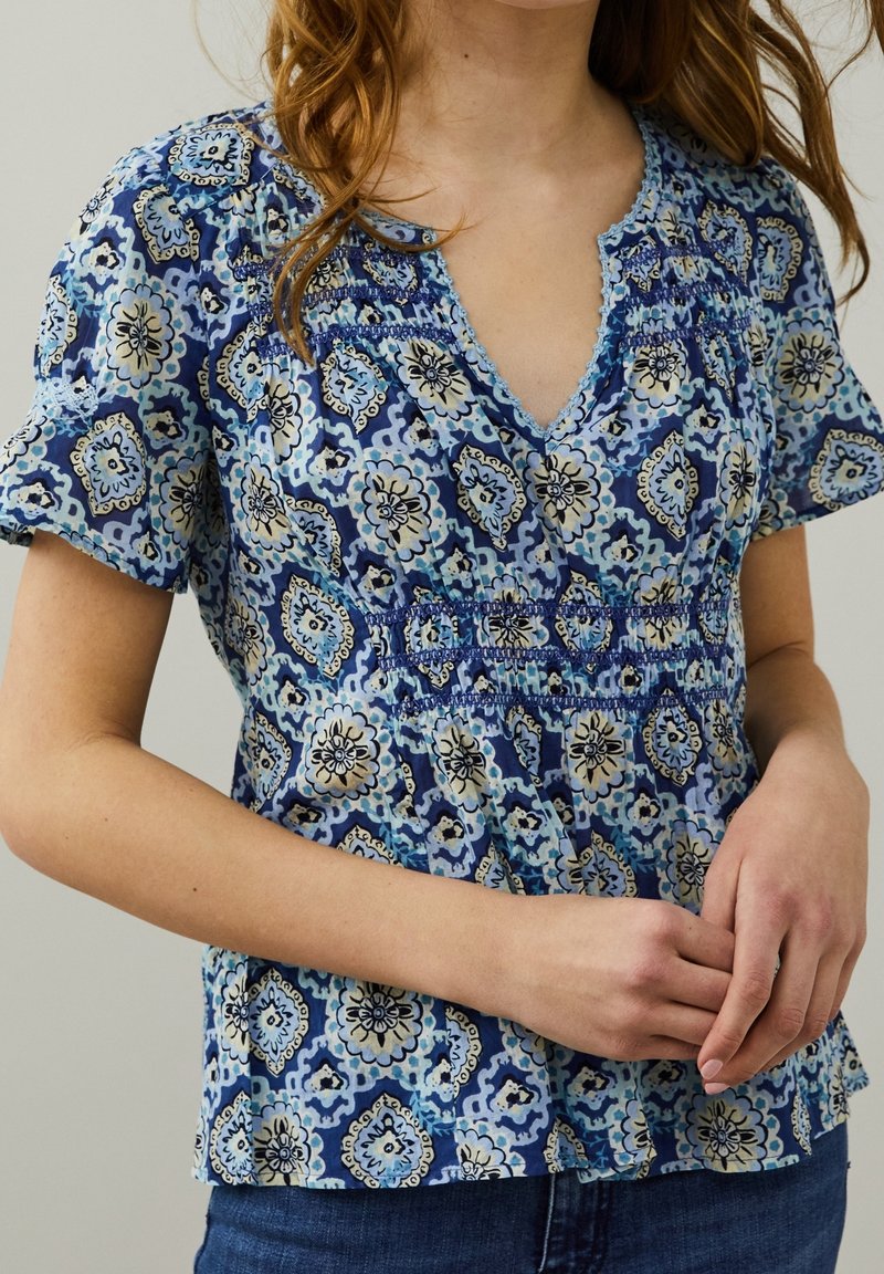 Blouse bleue à motifs avec un col en V, des manches courtes et un détail smocké à la taille ; présente des motifs floraux et géométriques.